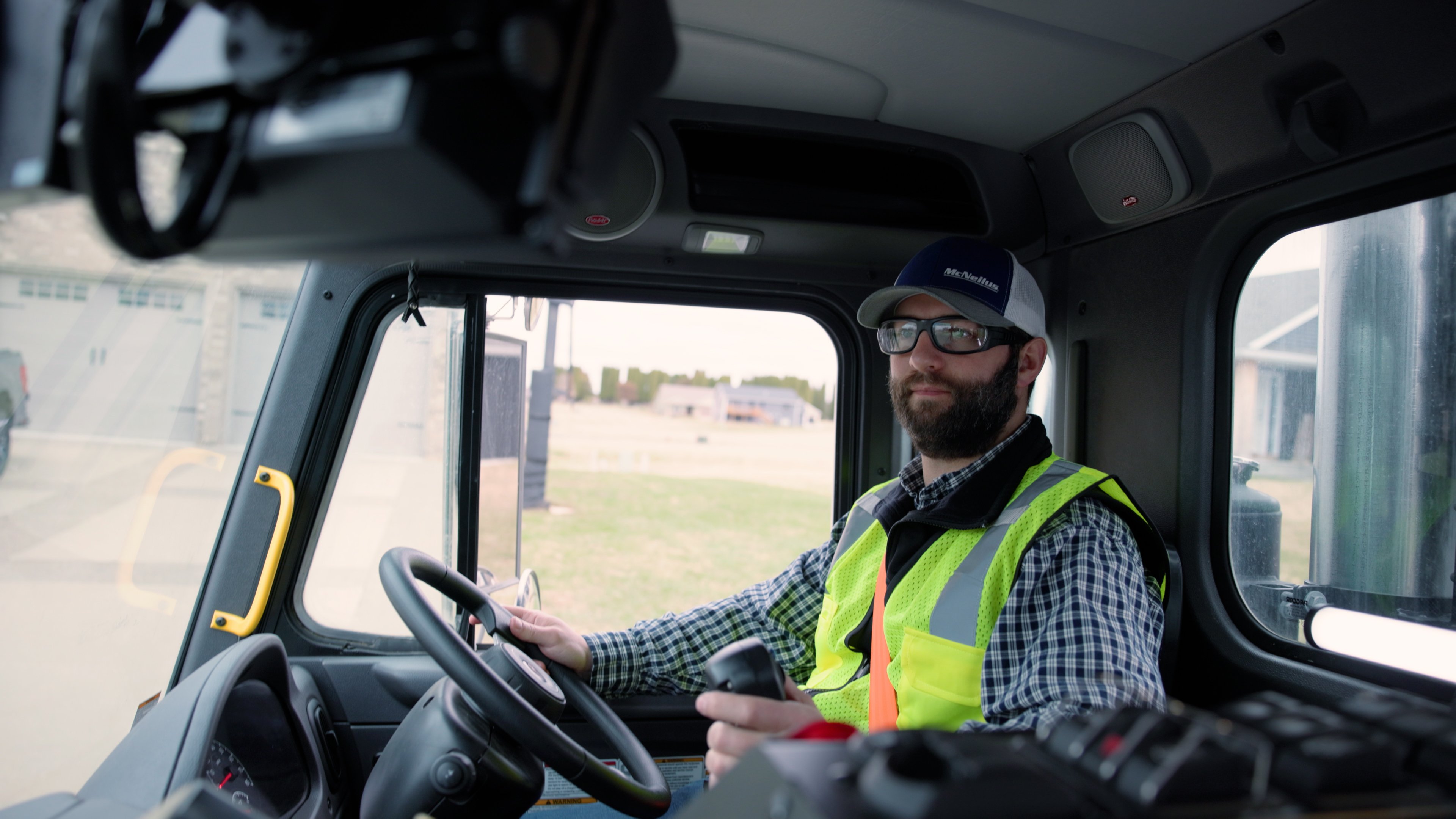 A garbage truck driver with a safety yellow vest on sitting in the driver's seat.