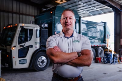 A man in a gray polo shirt stands in front of a Frontier garbage truck inside a garage.